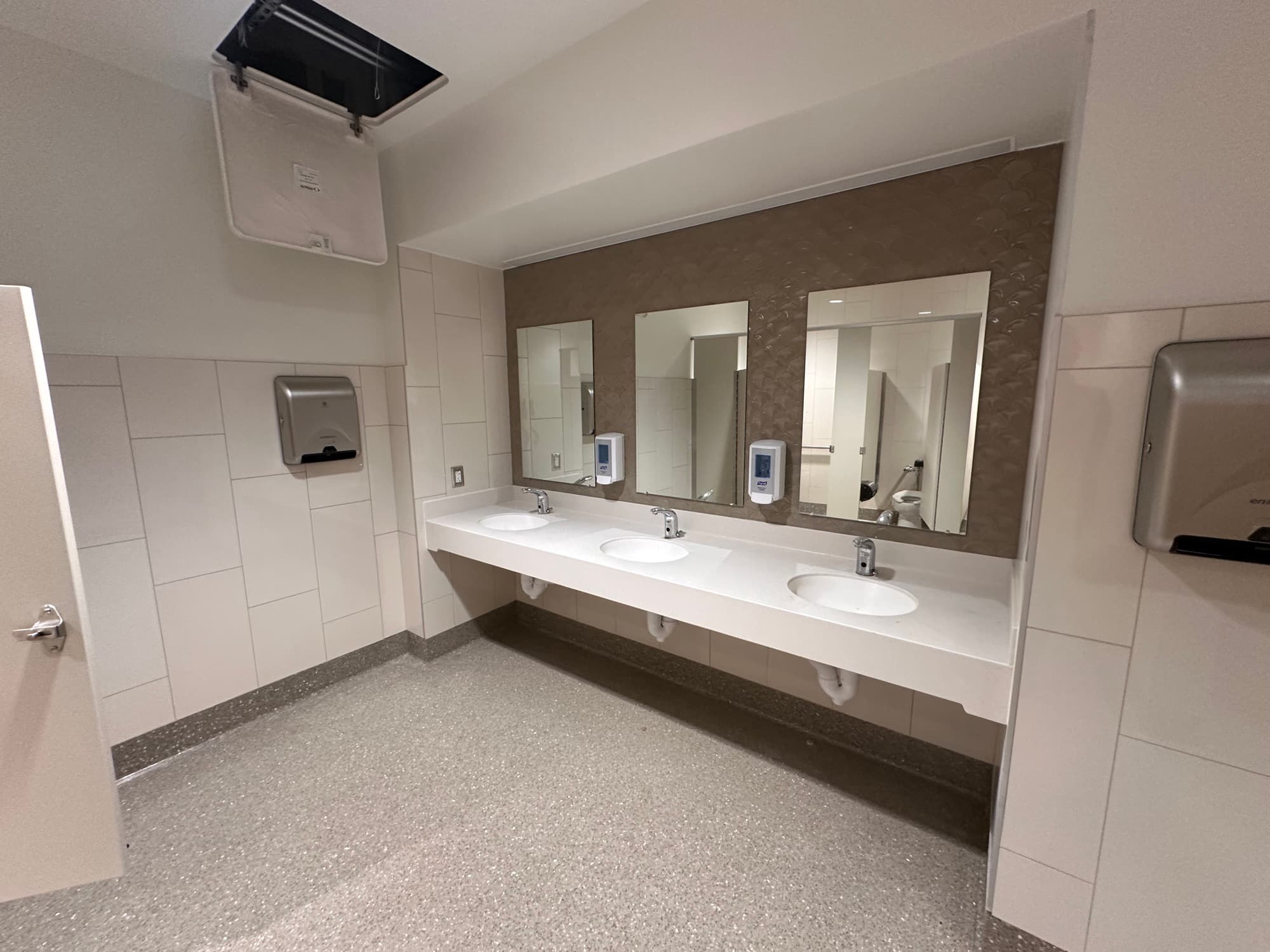 Completed commercial restroom with wall tile, continuous resilient flooring, and a three-sink vanity