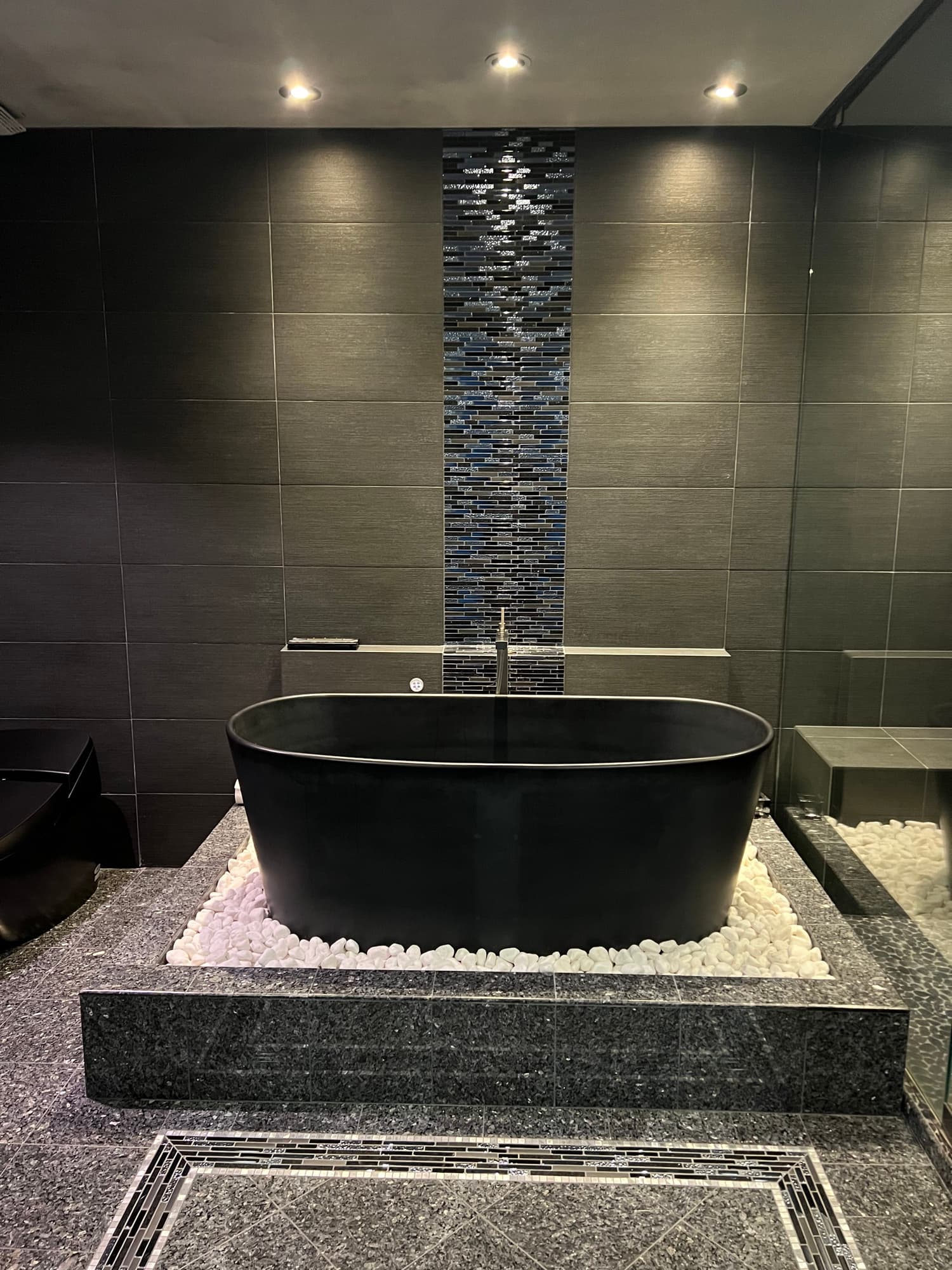 Completed dark tile bathroom with a freestanding tub, feature mosaic, and pebble details